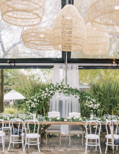 Elegant wedding reception setup with a long table, white chairs, greenery, and floral decorations under wicker pendant lights in a transparent tent.